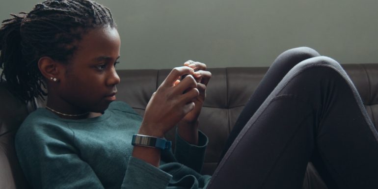 A young black girl laying on a couch, playing a game on her phone.